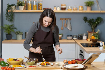 Young female in apron flavoring vegetable food with homemade sauce pesto while bending by kitchen table and plate with cooked stew