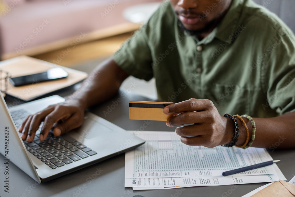 Poster hands of young man with credit card pressing keys of laptop keypad while sitting by table and shoppi