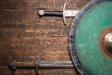 Fototapeta premium Two swords and the shield on the old medieval table flat lay concept background with copy space.