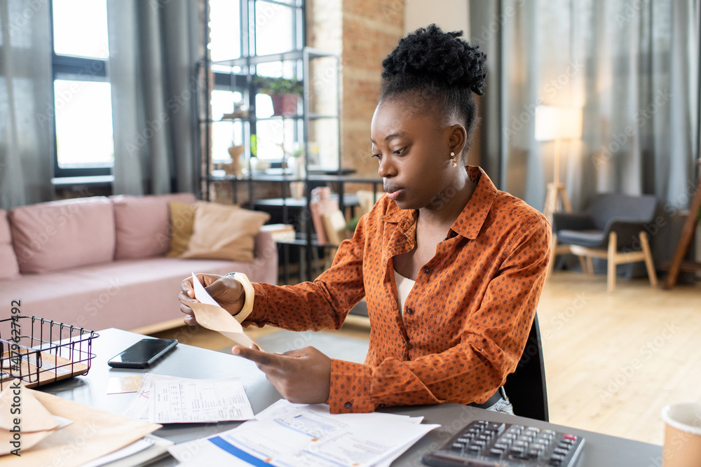 Poster young african woman sitting by table and putting filled form into envelope before sending it to orga