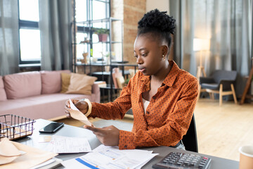 Young African woman sitting by table and putting filled form into envelope before sending it to...