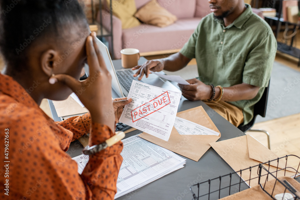 Poster Young stressed female looking at unpaid bill and touching her head while sitting by table in front of man networking