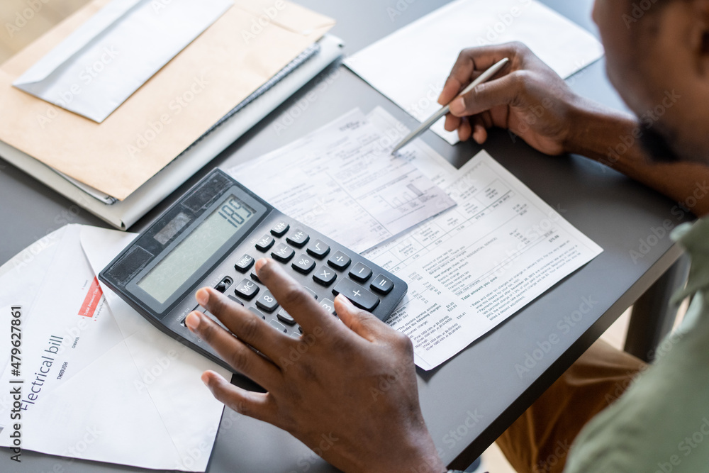 Aufkleber Hands of African man pressing buttons of calculator while checking data in financial papers and unpaid bills by table