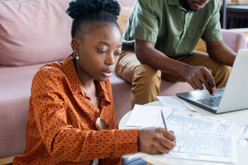 Young serious African woman making notes in financial document while sitting against man networking...