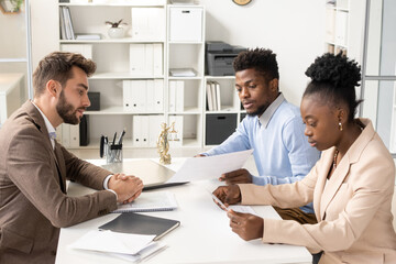 Young African woman in smart casualwear reading paper while her husband consulting with lawyer by table in office