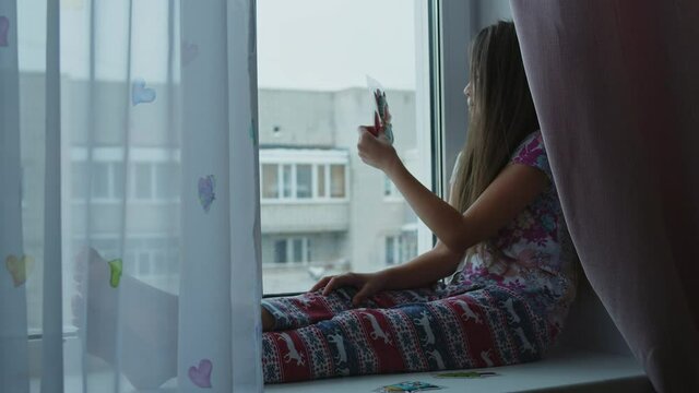 A little girl, sitting on the windowsill, decorates the window with colorful stickers.