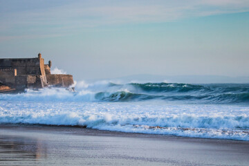 Large waves crash into a fortress near a beach. Carcavelos beach in Portugal