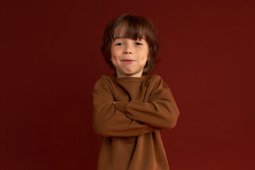 Charming shaggy charismatic joyful kid holding arms folded in bossy position, standing against red wall with cute smile on his lips, dressed in cashmere brown sweater. Carefree childhood