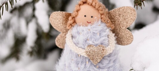 A soft textile doll with golden heart in hands sitting on the snow. Valentine’s Day, love, Merry Christmas, New Year concept. Banner.