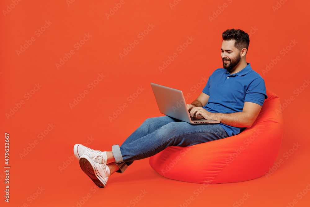 Wall mural Full body young smiling happy caucasian man wear basic blue t-shirt sit in bag chair hold use work on laptop pc computer isolated on plain orange background studio portrait. People lifestyle concept.