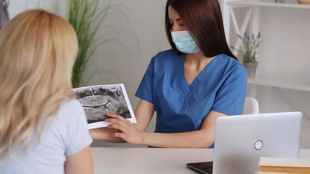 Dentist consultation. Female doctor. Medical therapy. Qualified woman protective mask showing teeth roentgenogram to patient in light room interior.