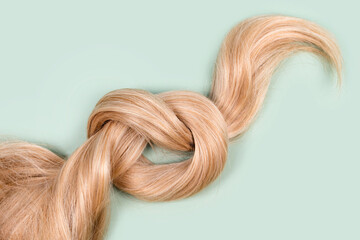 Blonde hair lock tied in knot. Strand of honey blonde hair on mint background, top view. Hairdresser service, hair strength, haircut, hairstyle, dying or coloring, hair extension, treatment concept. - obrazy, fototapety, plakaty