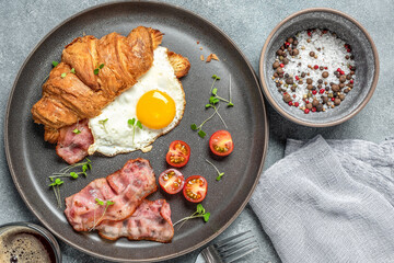 Croissant sandwich with fried egg, bacon, tomato, microgreens, coffee. Delicious appetizing breakfast. Gray grunge background.Top view, flat lay