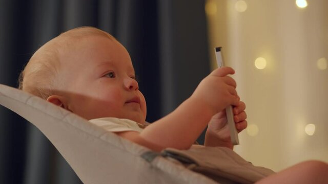 Child watching TV in living room, cute baby boy holding tv remote sitting in rocking chair. High quality 4k footage