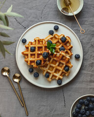 Waffles with addition of fresh blueberries and honey on a white plate on table with natural grey linen tablecloth close up view action shot girl pouring honey on waffles