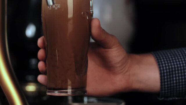 Barman in plaid shirt pours a pint of cold dark beer into a clear glass, the foam slowly settles. High quality 4k footage