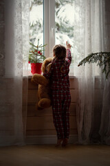 A cute little girl in New Year's pajamas with a teddy bear looks out the window. Merry Christmas and happy New Year