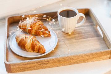 Freshly baked croissant on a gray round plate, white cup with coffee and garland on a tray on the table