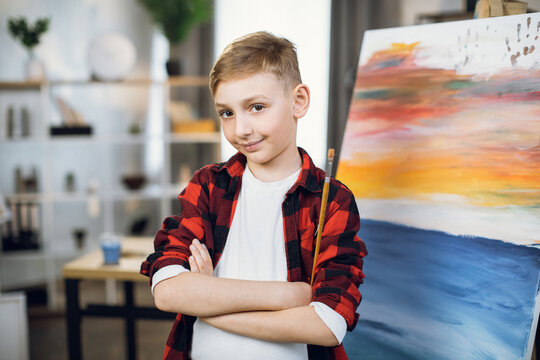 Happy caucasian boy in casual wear posing with crossed arms near his masterpiece on easel. Talented child creating abstract pattern with brush and paint in studio.