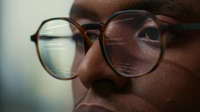 Vfx of running programming code reflected in software developer glasses. Extreme closeup of african american programer working focused looking at computer screen while typing.