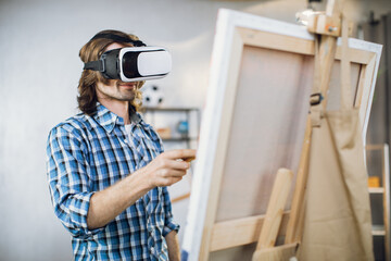 Inspired young man drawing with brush on easel while wearing modern VR goggles. Innovative technology for art. People and hobby concept.