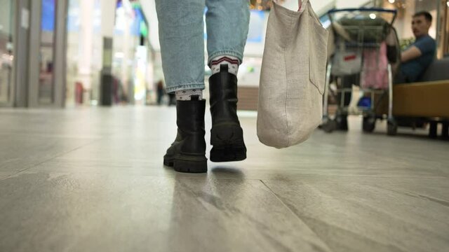 Young slim woman wearing jeans socks and boots walks on white tiled floor near clothes shops in shopping centre close view