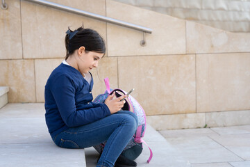 Little girl with cellphone sitting on stairs by school backpack. Young kid using smart phone. Childhood technology addiction concept