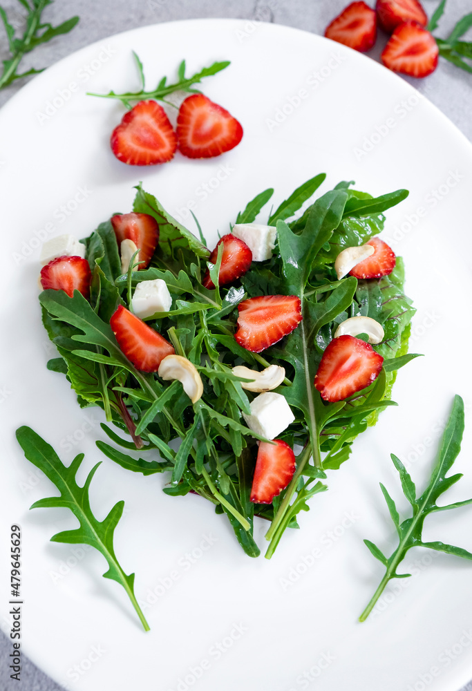 Wall mural Delicious salad of strawberry with mix of herbs, arugula, spinach leaves, feta cheese and cashew nuts in heart shape on white plate. Vegetarian food and raw food diet.