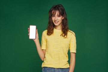 Smiling beautiful young girl show phone with blank mock-up screen, advertising new apps on green studio background