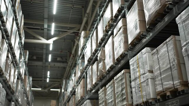 Rows of high metal storage racks with boxes of stuff items standing on shelves and wrapped in polyethylene in big warehouse of mall