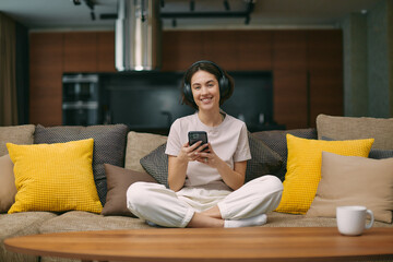 Obraz premium Smiling young girl in headphones holding phone, using mobile app, listening to music, learning foreign language on couch
