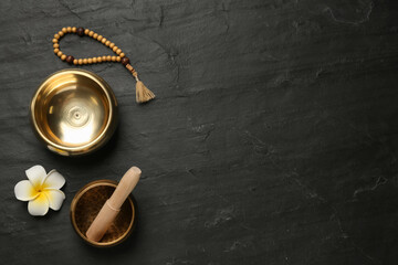 Golden singing bowls with mallet, beads and flower on black table, flat lay. Space for text