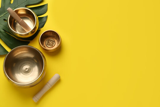 Golden singing bowls, mallets and monstera leaf on yellow background, flat lay. Space for text