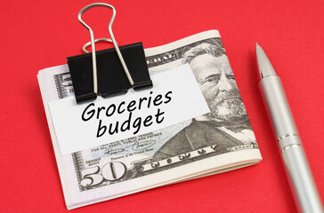 On a red background lies a pen and dollars clamped with a clip with the inscription on paper - Groceries budget