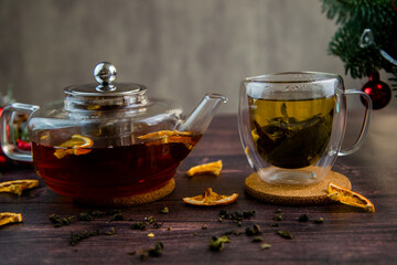 A transparent teapot and a cup of tea stand against a background of dark wood, slices of dried orange and Christmas decorations. High quality photo