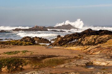 Scenic landscape of the Atlantic Ocean coast with beating waves against old brown stones with an old lighthouse and high splashes of blue water with white foam and clear sky, Portugal