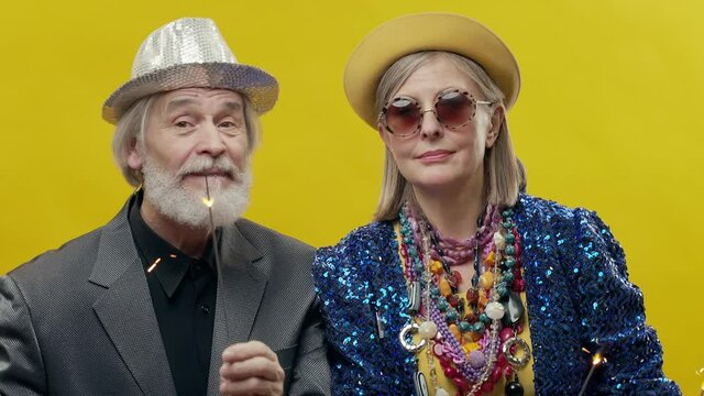 Elderly Man And Woman. Stylish Elderly Woman On Yellow Background. She Is Wearing Hat And Glasses. Stylish Man With Beard In Hat. They Have Sparklers In Their Hands.