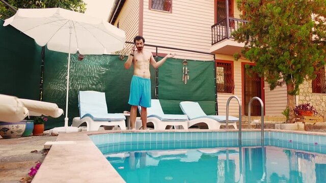 Young man in swimwear is standing near swimming pool on villa and talking on smartphone gesturing actively. Caucasian bearded tourist solves work problems distantly during his vacations. Holiday theme