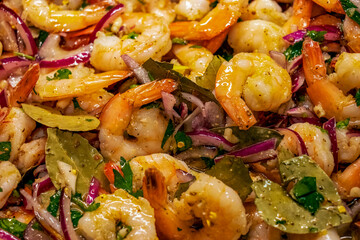 Close-up of cooked shrimp with tails on in a marinade of olive oil and lemon juice with whole bay leaves, thin sliced red onion, crushed red pepper, garlic, and fresh parsley.