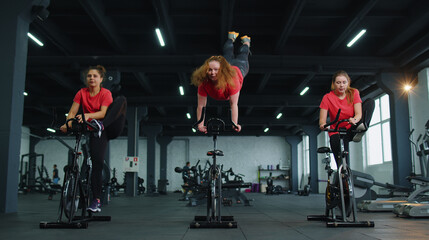 Class group of young women exercising on stationary cycle machine bike in gym. Modern sport activity, workout, healthy lifestyle. Athletic girls spin bike, making tricks indoors. Slow motion