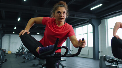 Group of athletic young girls training stretching on spin stationary bike riding in gym. Fit women performs aerobic training workout cardio routine on simulators cycle training on exercise bike indoor