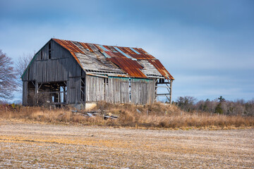 An old barn with weathered gray boards and a rusty roof sits in a farm field. © Ken