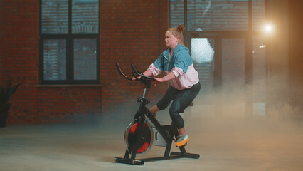 Caucasian woman coach spin on stationary bike in smoky gym. Athletic girl performing aerobic riding training exercises on cycle bike. Weight Loss Cardio Healthy Lifestyle. Modern sport indoors - obrazy, fototapety, plakaty