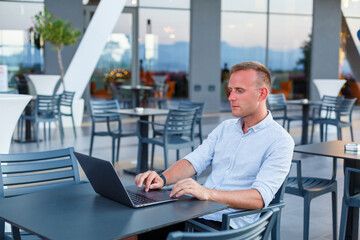 Naklejka na ściany i meble Successful young executive looking while sitting at a table with a laptop on a hotel terrace and finding time for remote control of employees while on vacation in an exotic resort