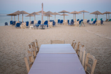 Empty white table with table cloth and chairs on sand in typical Greek tavern restaurant at the sea in Nea Skioni fishing village with empty umbrellas and deck chairs in background. Nea Skioni,Greece