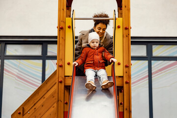 A young woman, nanny, teaching a little boy how to ride slides at playground.