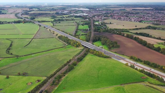 Aerial footage of the M62 Motorway located in Leeds showing a train track going under the motorway and a train traveling on the tracks along side farmers fields and beautiful scenic area in Yorkshire
