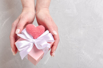 Top view of a woman's hand holding a pink gift box with a white ribbon and a knitted pink heart. A Valentine's Day card. Space for copying. Flat position, top view.