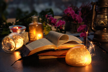 Magic lights with sparkles and orange glow in various glass jars. Wintertime with lights and old books. Stack of old vintage books with one open. Magenta orchid flowers. Romantic indoor background.