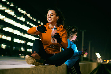 A happy female runner taking a break. A happy night runner sitting on the pavement, drinking water...